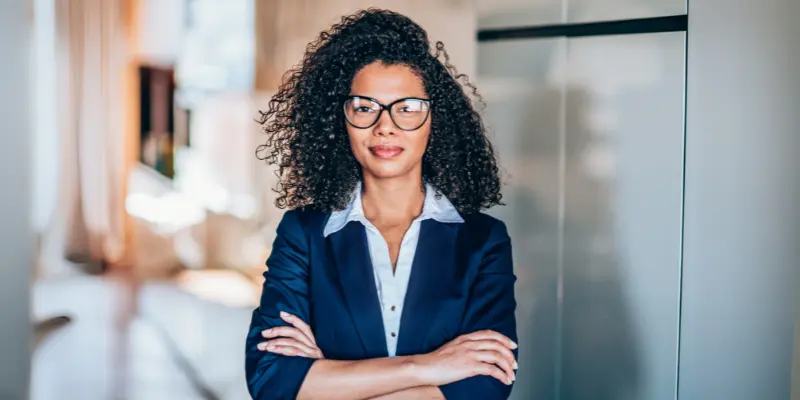A professional woman with curly hair, wearing a navy blazer and white shirt, stands confidently in an office.