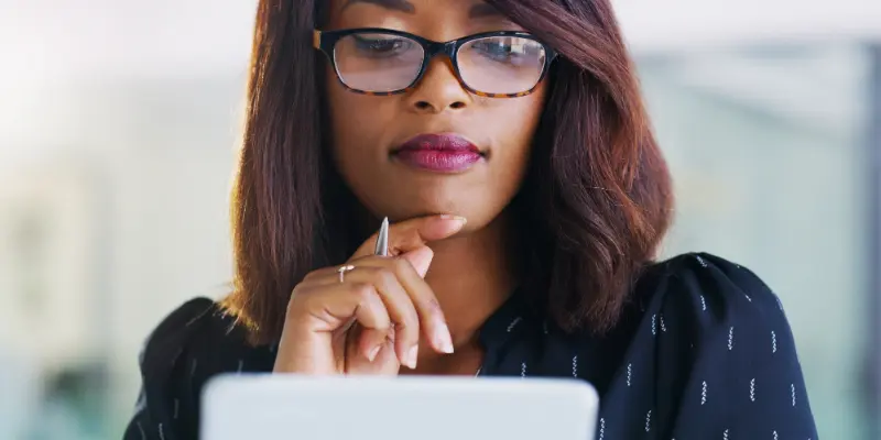 A person with shoulder-length hair, wearing a dark blouse, holding a pen to the chin while looking at a tablet.